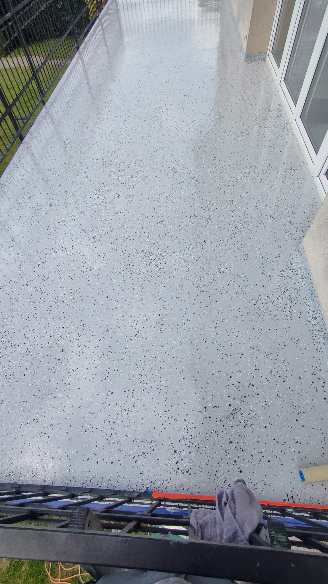 Balkon z szarą, błyszczącą posadzką żywiczną z czarnymi i białymi drobinkami, widok z góry, z fragmentem balustrady i okna w tle.