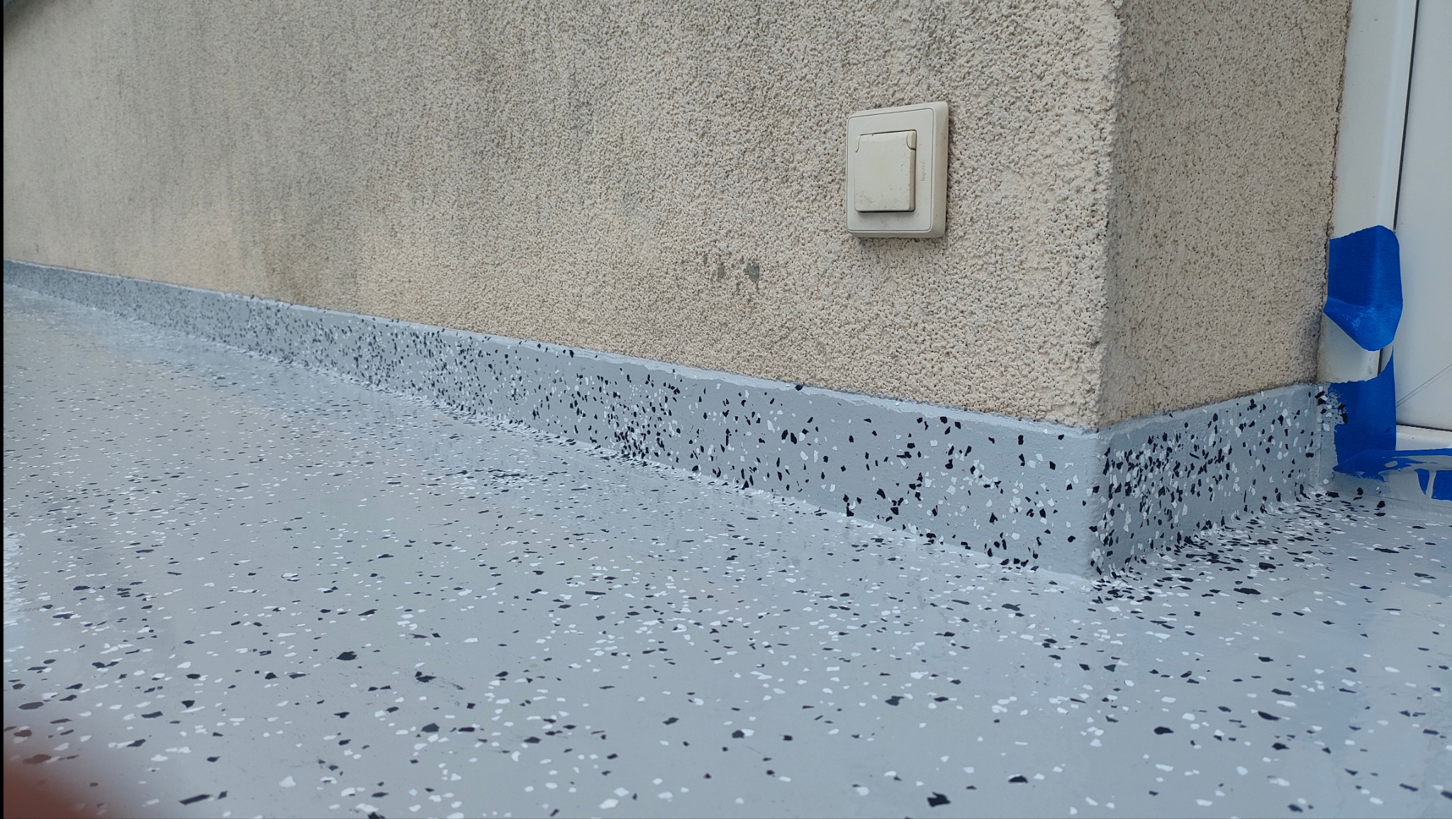 Szara, żywiczna posadzka z czarnymi i białymi elementami dekoracyjnymi, wykończona przy ścianie, z widocznym gniazdkiem i zabezpieczeniem folią malarską.