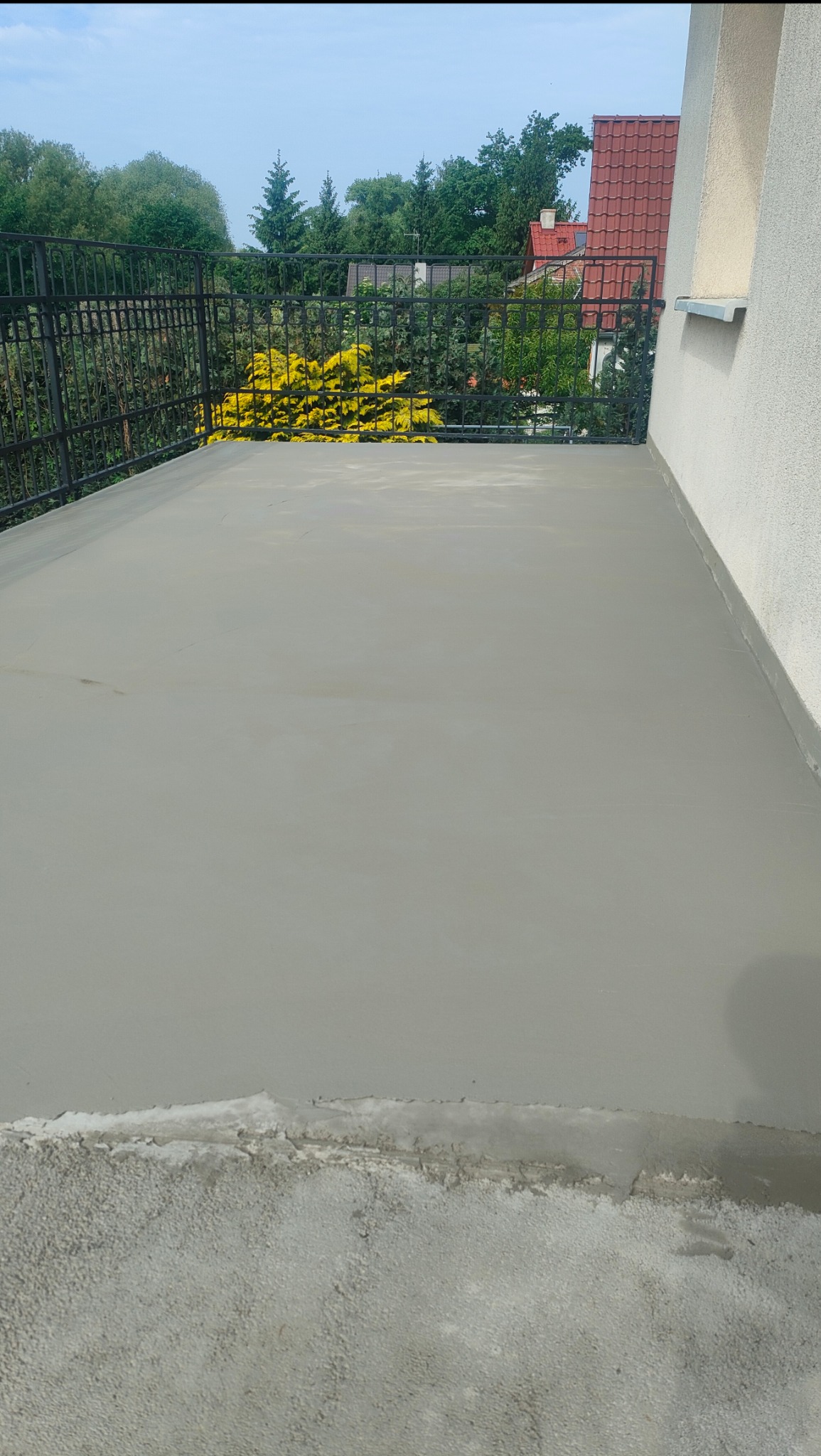 Wyrównana, szara wylewka na balkonie z metalową balustradą i widokiem na zieleń w tle. Świeżo położona, gładka powierzchnia przygotowana pod dalsze prace wykończeniowe.
