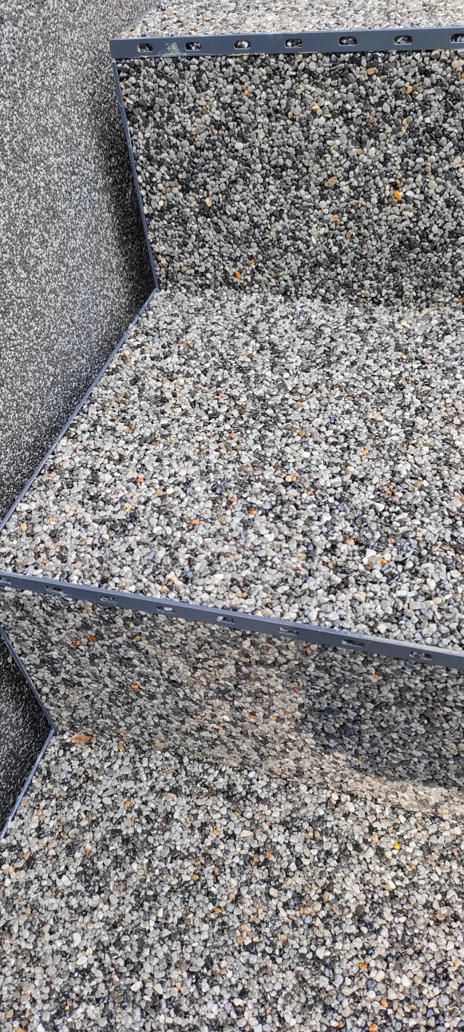 Wykończenie schodów żywicą epoksydową z kamyczkami, widoczne krawędzie schodów zabezpieczone listwami. Szara kolorystyka, drobna faktura kamieni.