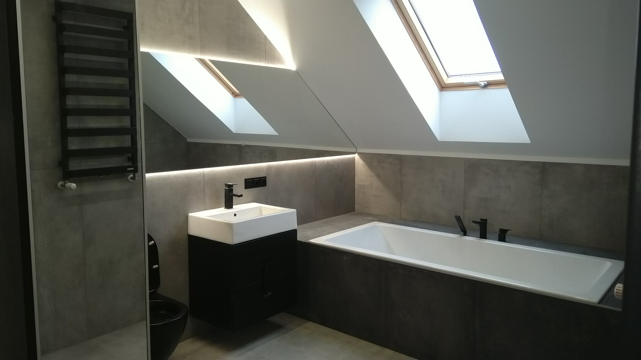 Nowoczesna łazienka na poddaszu z oknem dachowym, szarą glazurą, białą wanną i umywalką na czarnej szafce, podświetlona listwą LED.