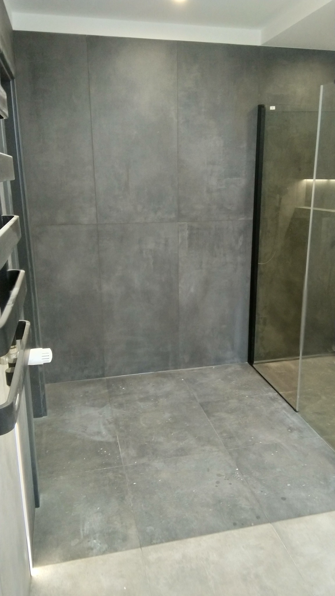 Szare, matowe płytki ceramiczne na ścianach i podłodze w łazience z kabiną prysznicową z czarnymi profilami, widoczny fragment grzejnika drabinkowego.