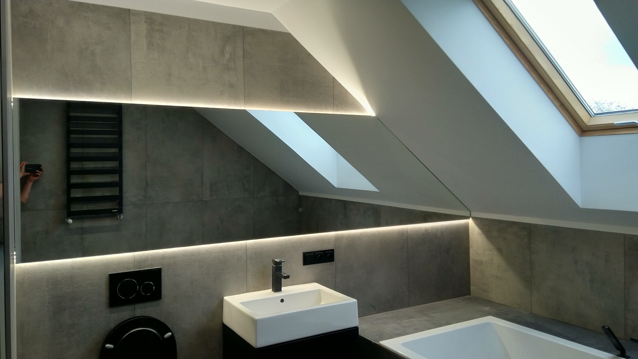 Nowoczesna łazienka na poddaszu z szarymi płytkami imitującymi beton, podświetlonym lustrem LED, białą umywalką i wanną oraz oknem dachowym.