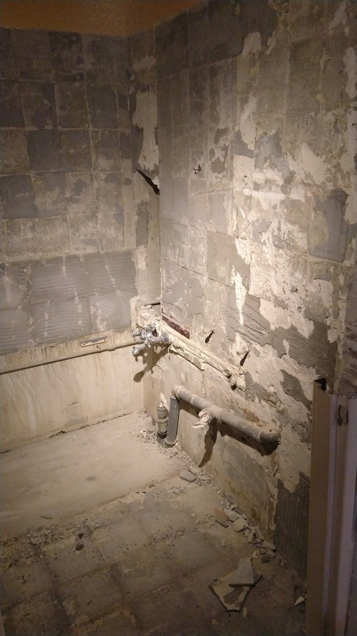 Wnętrze łazienki w trakcie demontażu płytek, widoczne odsłonięte rury, ślady po kleju i gruz na podłodze.