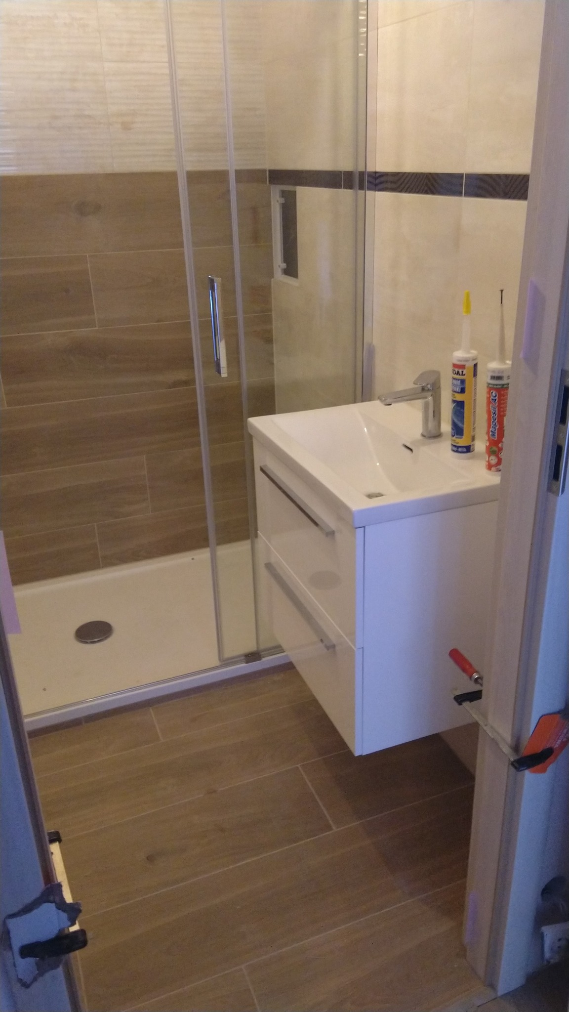 Nowoczesna łazienka w trakcie wykańczania: widoczna kabina prysznicowa, biała szafka z umywalką, brązowe płytki imitujące drewno na podłodze i ścianach, oraz tuby z silikonem i ścisk stolarski przy...