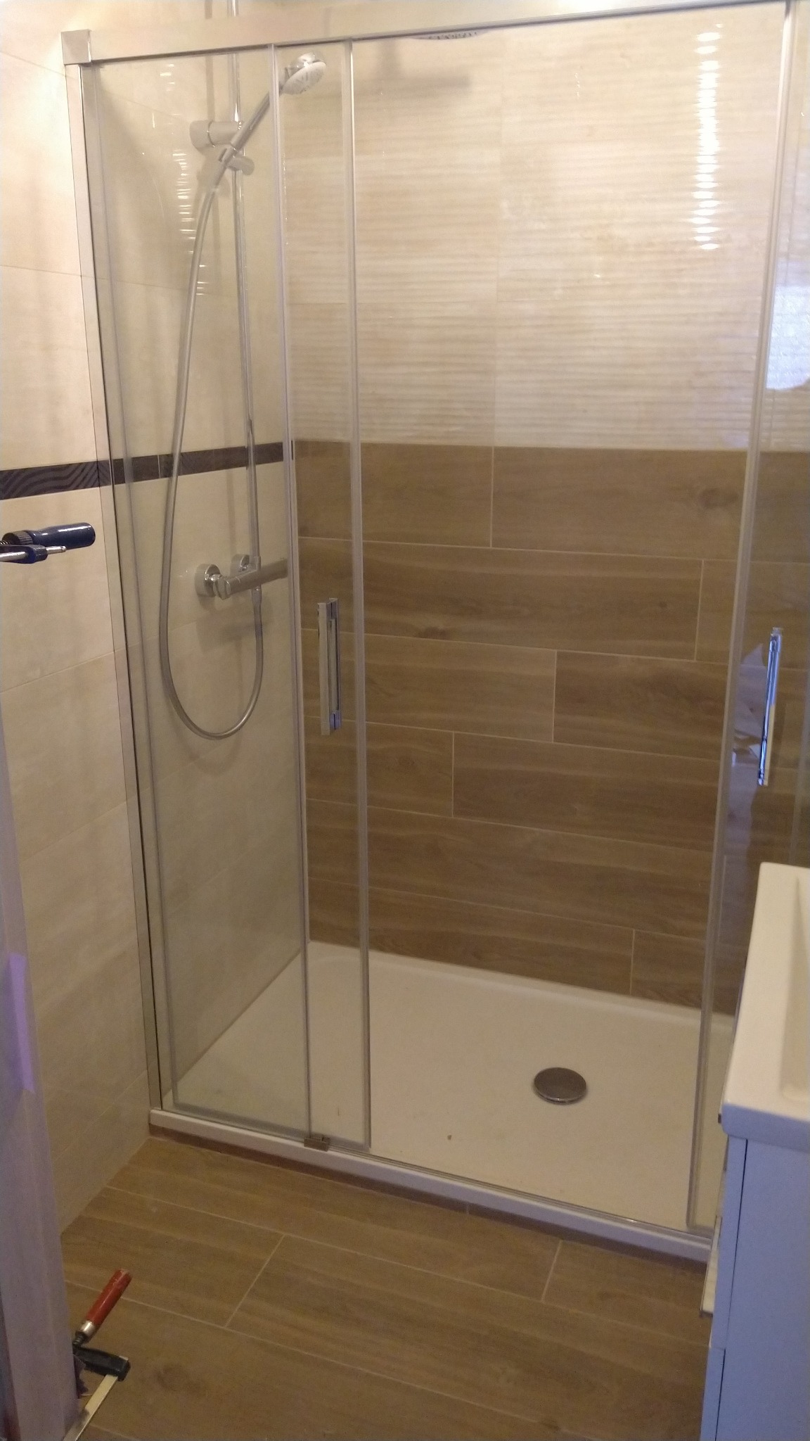 Kabina prysznicowa z przesuwnymi drzwiami, zamontowana na białym brodziku, z widoczną armaturą i dwukolorową glazurą w tle; na podłodze płytki imitujące drewno, w rogu widoczne narzędzia.