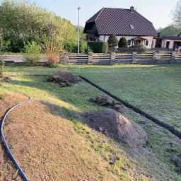 Wykopany rów w trawniku z widocznymi pryzmami ziemi i niebieskim wężem, przygotowanie do instalacji w ogrodzie.