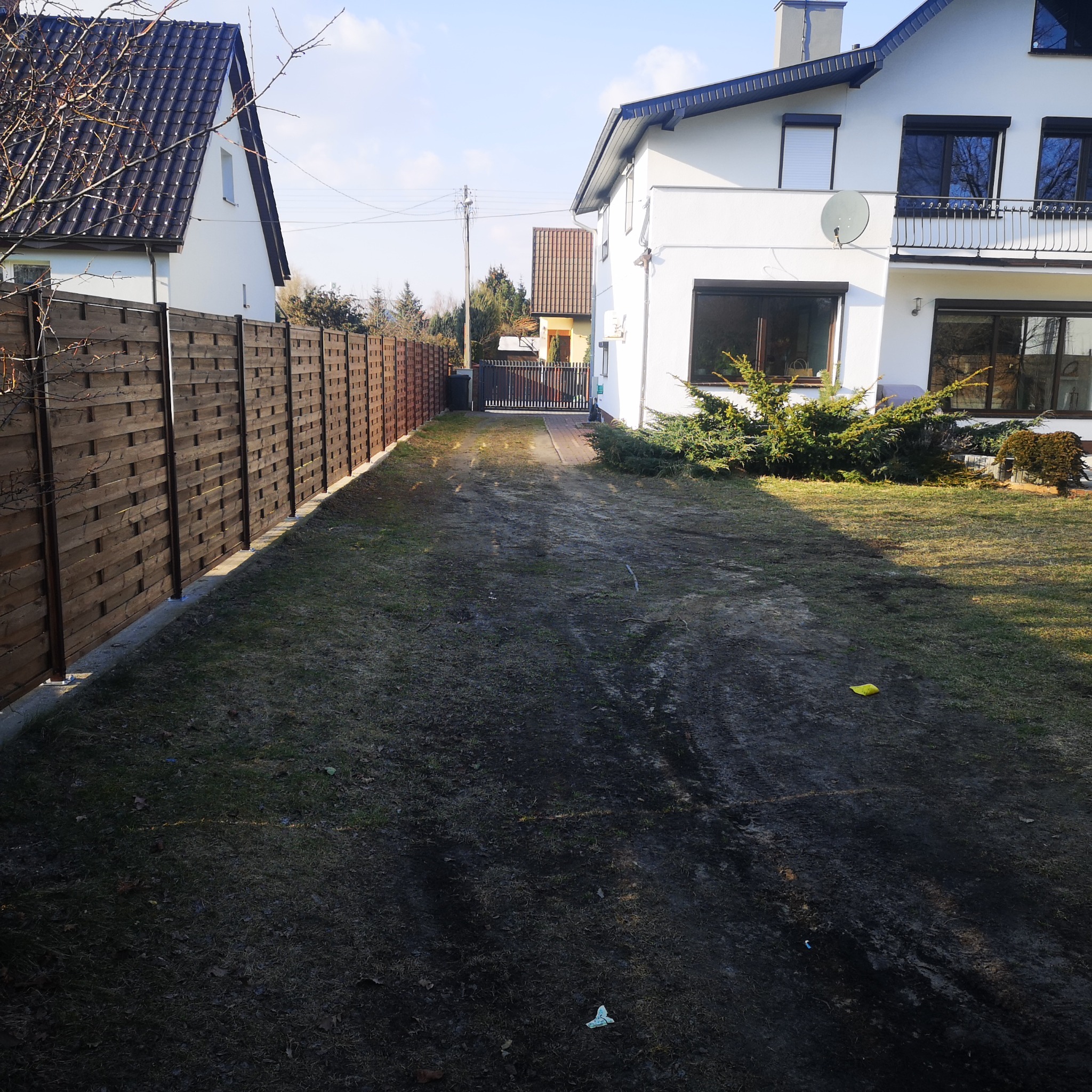 Długa, nowa, drewniana, pozioma, pleciona, ciemnobrązowa deska ogrodzeniowa oddzielająca posesję od sąsiada, z widocznym podłożem trawiastym przed i za ogrodzeniem. W tle dom z bramą wjazdową.