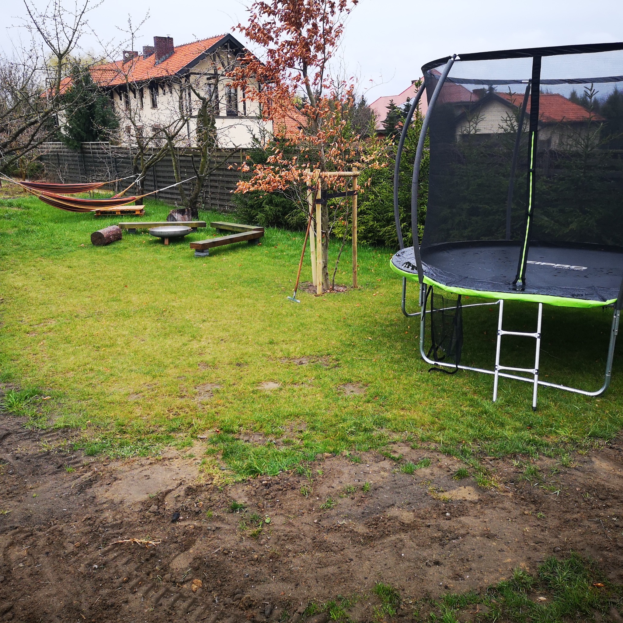 Ogród z trampoliną, hamakiem i świeżo posadzonym drzewkiem podpartym drewnianymi stemplami, widoczne ślady po pracach ziemnych.