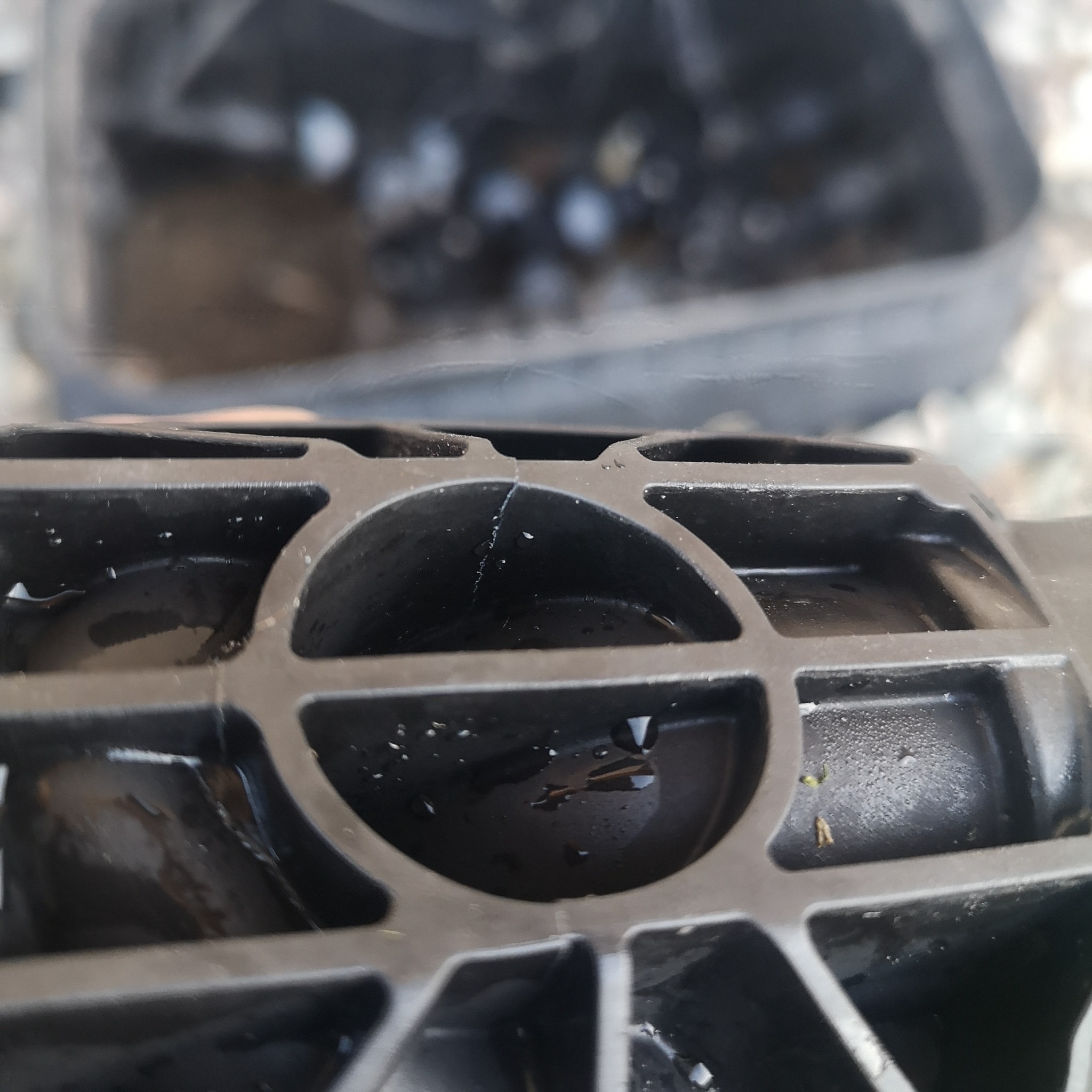 Czarny element systemu nawadniania z widocznym pęknięciem i kroplami wody, z niewyraźnym tłem żwirowym.