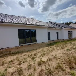 Parterowy dom z białymi ścianami i ciemną dachówką, widoczne okna i drzwi, teren wokół domu jeszcze niezagospodarowany, pochmurne niebo.