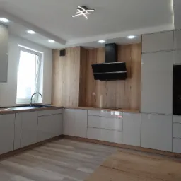 Nowoczesna kuchnia z szarymi, lakierowanymi frontami, drewnianym blatem i okładziną ściany, czarnym okapem i piekarnikiem w zabudowie, oświetlona lampą sufitową w kształcie gwiazdy.