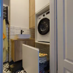 Nowoczesna łazienka z zabudowaną pralką, umywalką nablatową na drewnianej szafce i wzorzystą podłogą, widok zza uchylonych drzwi.