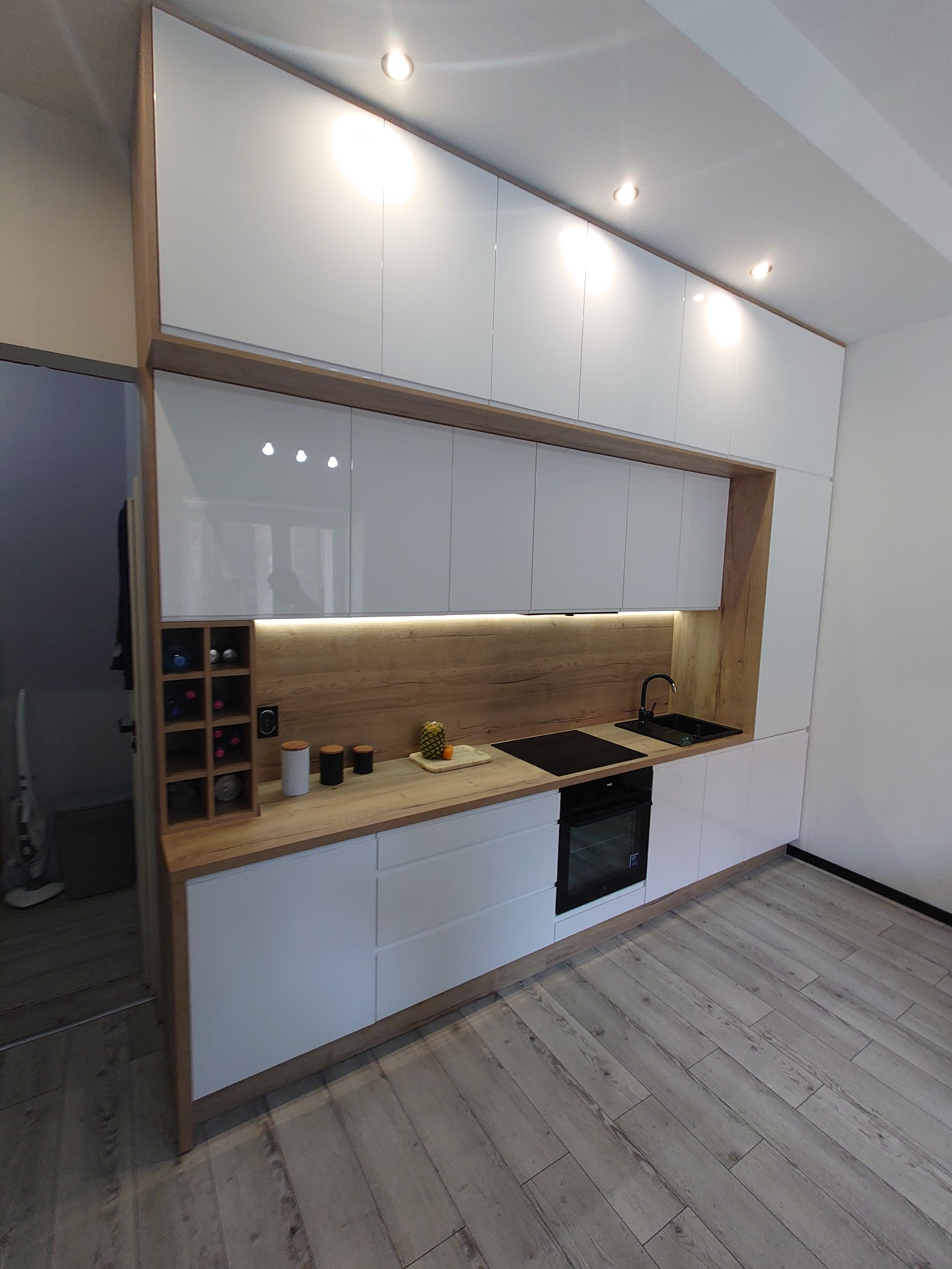 Nowoczesna kuchnia z białymi, błyszczącymi szafkami, drewnianym blatem i podświetleniem LED, z widocznym regałem na wino i elementami dekoracyjnymi.