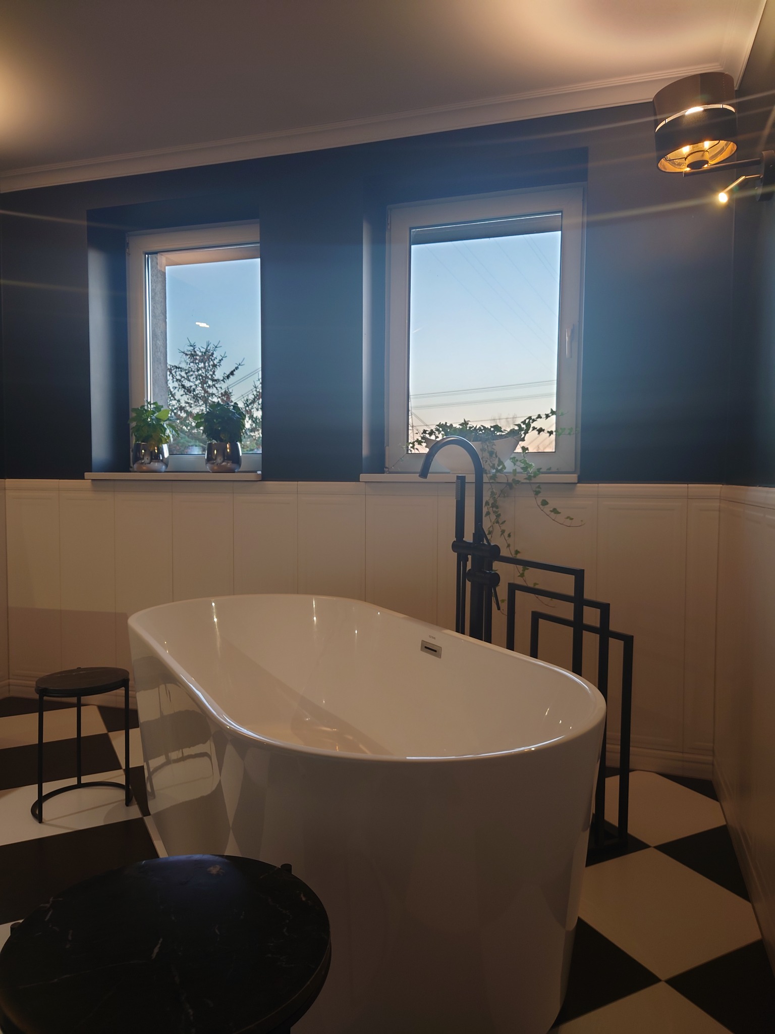 Nowoczesna aranżacja łazienki w Płocku z wolnostojącą białą wanną, czarną armaturą i czarno-białą szachownicą na podłodze, doświetlona przez dwa okna z roślinami.