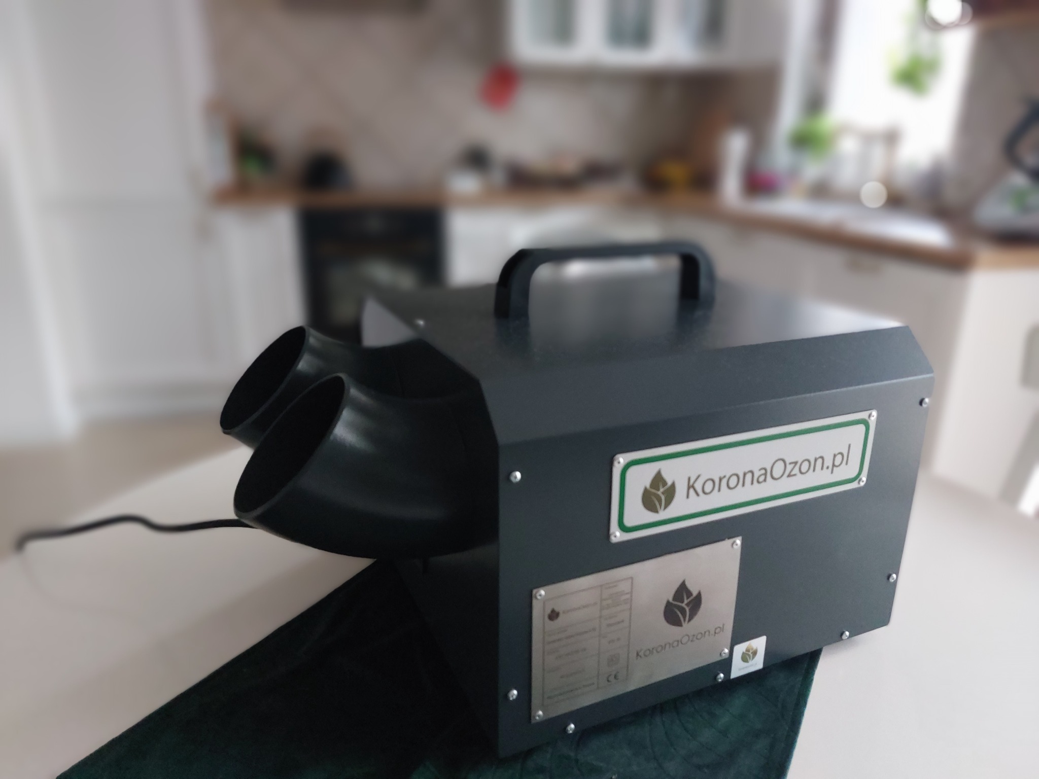 Ozonator przemysłowy marki KoronaOzon.pl, czarna obudowa z dwoma wylotami powietrza, umieszczony na blacie w kuchni, widoczne logo firmy na obudowie.