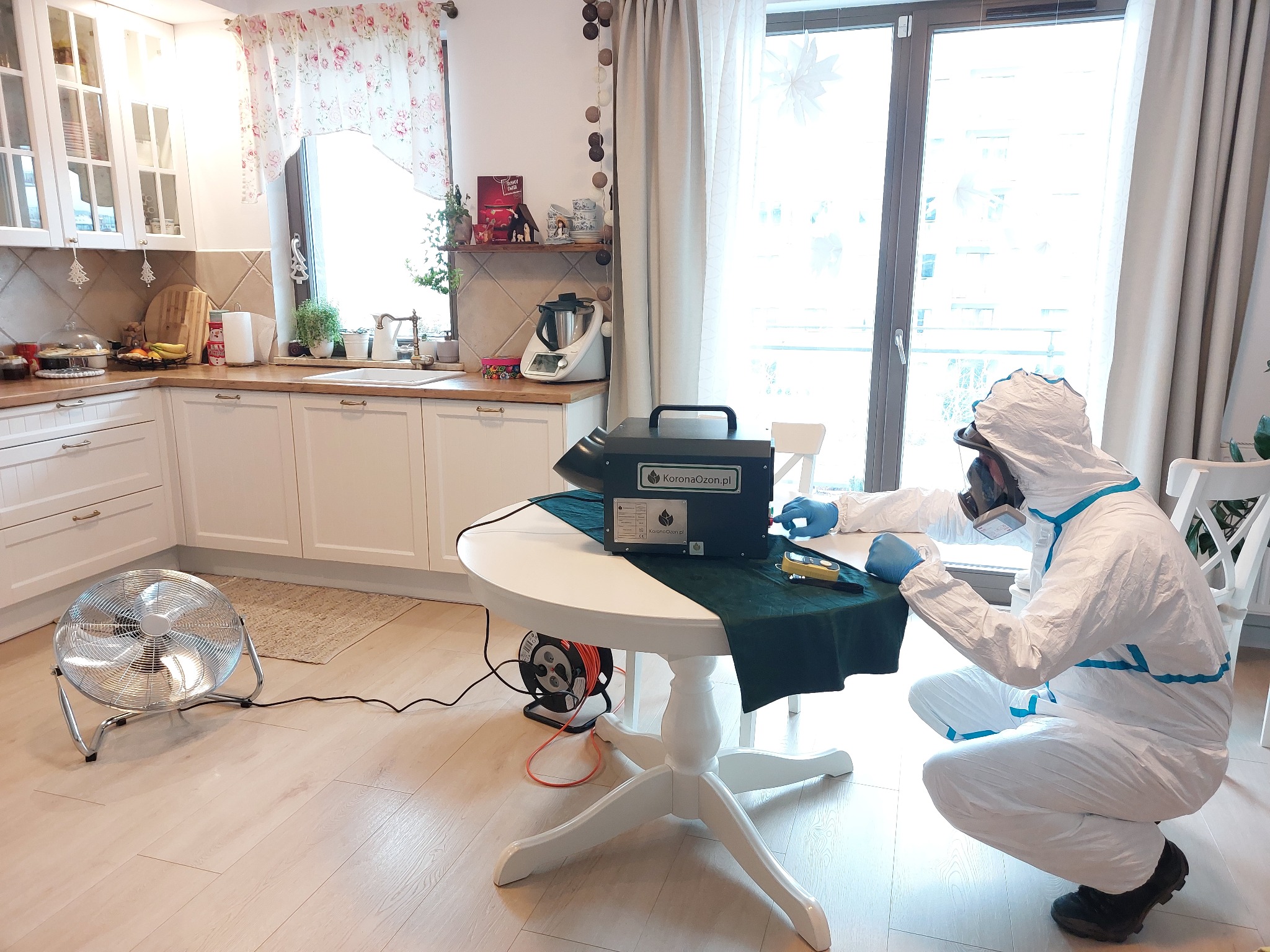 Osoba w kombinezonie ochronnym i masce, w trakcie ozonowania pomieszczenia mieszkalnego, z widocznym ozonatorem na stole i wentylatorem podłogowym.