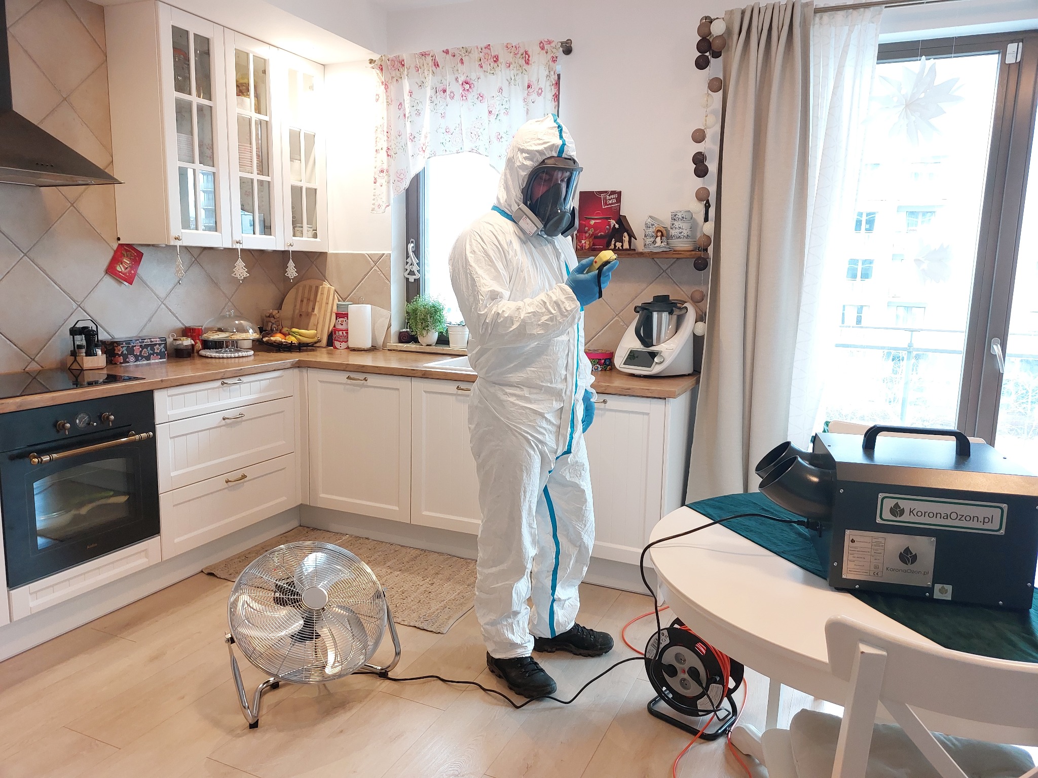 Pracownik w kombinezonie ochronnym i masce, trzymający w rękawicach mały przedmiot, w trakcie pracy z ozonatorem KoronaOzon.pl w kuchni w Gdańsku, z widocznym wentylatorem i przedłużaczem na podłodze.