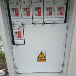 Otwarta skrzynka elektryczna z pięcioma bezpiecznikami o numerach 605, 628, 629, 609, 610 i jednym bezpiecznikiem o numerze 511 wewnątrz, widoczny znak ostrzegawczy o napięciu elektrycznym.