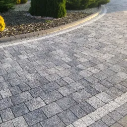 Ułożona szara kostka granitowa tworząca ścieżkę z obrzeżem, widoczne rośliny ozdobne w tle.
