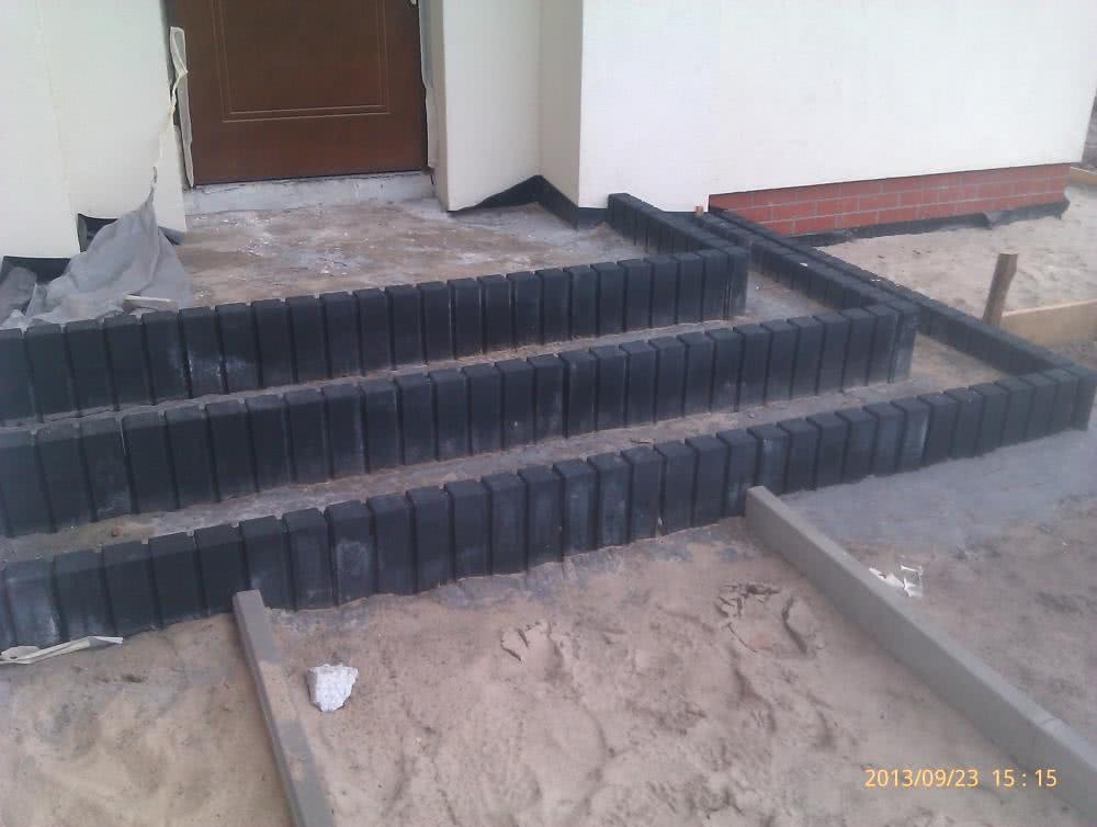 Wstępny etap budowy schodów wejściowych z czarnej kostki brukowej, widoczne przygotowane podłoże z piasku i częściowo ułożone obrzeża.