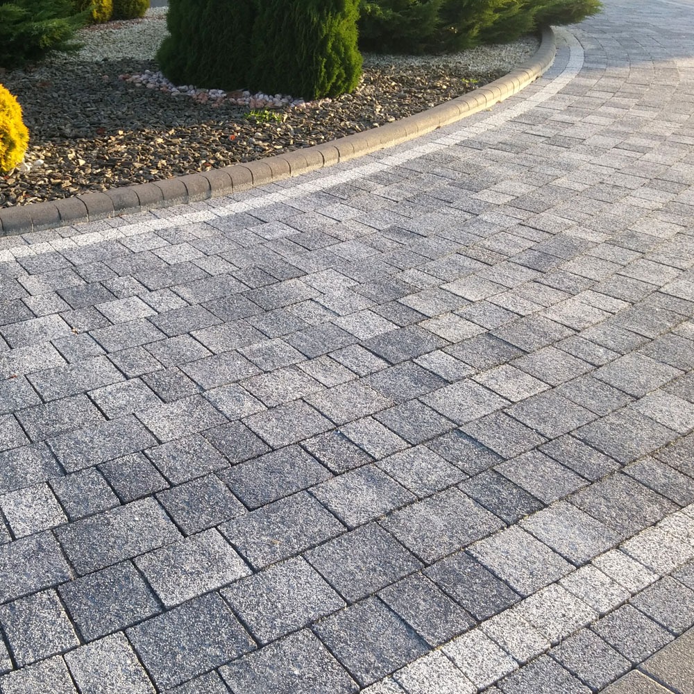 Ułożona szara kostka granitowa tworząca ścieżkę z obrzeżem, widoczne rośliny ozdobne w tle.