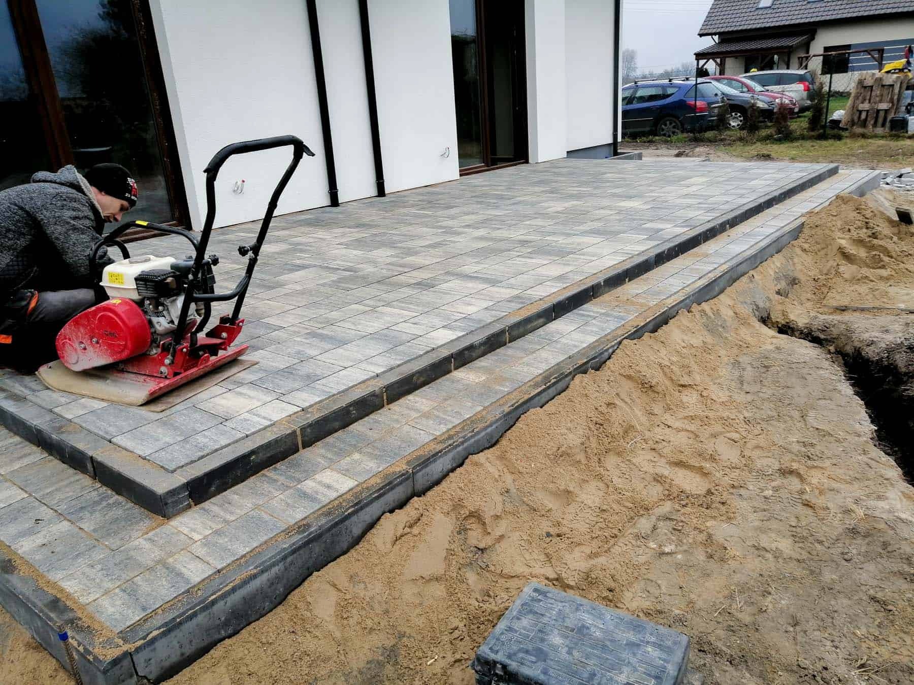 Prace brukarskie w toku: zagęszczarka wibrująca ubija szaro-beżową kostkę brukową na tarasie przed nowoczesnym domem, widoczne obrzeża i piasek przygotowany pod kolejne etapy.