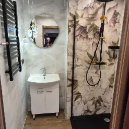 Nowoczesna łazienka z kabiną prysznicową typu walk-in, umywalką z szafką, grzejnikiem drabinkowym i lustrem o nieregularnym kształcie. Ściany wyłożone płytkami imitującymi marmur oraz płytkami...