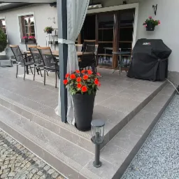 Wykończony taras z szarych płytek, schodami i obrzeżem z kostki. Na tarasie stół z krzesłami, grill i kwiaty w donicach. Oświetlenie tarasowe.