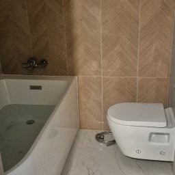 Wykończona łazienka z białą wanną wypełnioną wodą, podwieszaną toaletą i brązowymi płytkami ułożonymi w jodełkę na ścianie oraz marmurową podłogą.