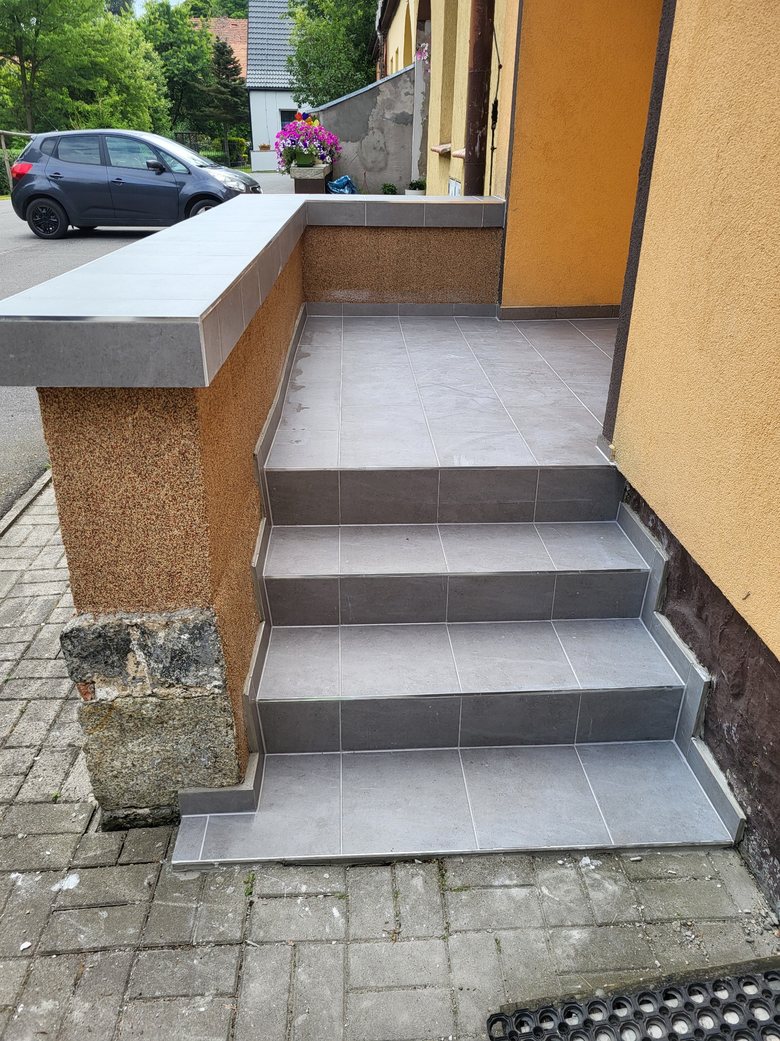 Wykończone schody wejściowe do domu z szarych płytek ceramicznych, widoczna elewacja w kolorze musztardowym i fragment samochodu w tle.