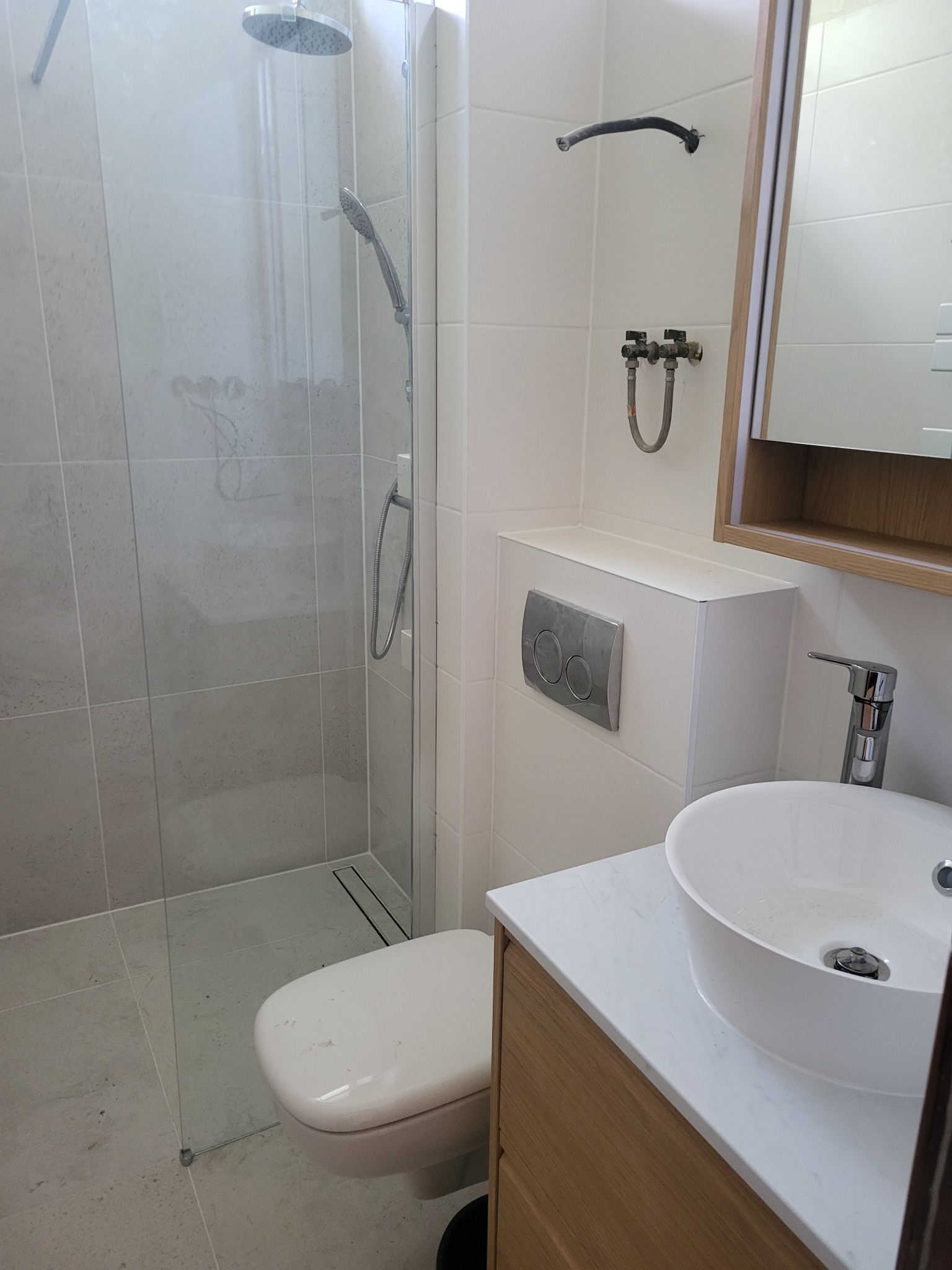 Nowoczesna łazienka z kabiną prysznicową typu walk-in, podwieszaną toaletą z chromowanym przyciskiem i umywalką nablatową na szafce z drewnianym fornirem.