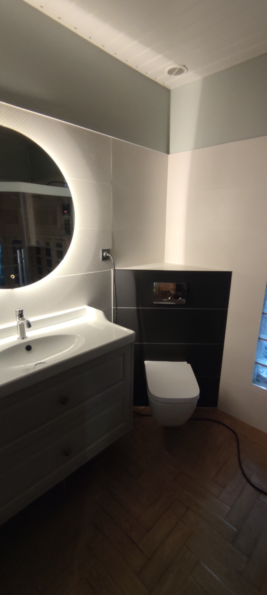 Nowoczesna łazienka z okrągłym lustrem podświetlanym LED, białą umywalką z szafką, wiszącą toaletą i czarnymi płytkami w strefie WC.