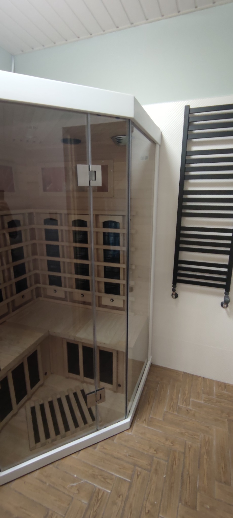 Zamknięta kabina sauny infrared z drewnianym wnętrzem i szklanymi drzwiami, widoczna w rogu łazienki obok czarnego grzejnika drabinkowego.