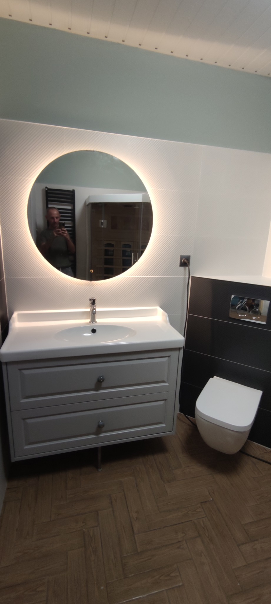 Nowoczesna łazienka z okrągłym lustrem podświetlanym LED, szarą szafką umywalkową z dwoma szufladami, wiszącą toaletą i parkietem ułożonym we wzór jodełki; w lustrze odbija się mężczyzna robiący...