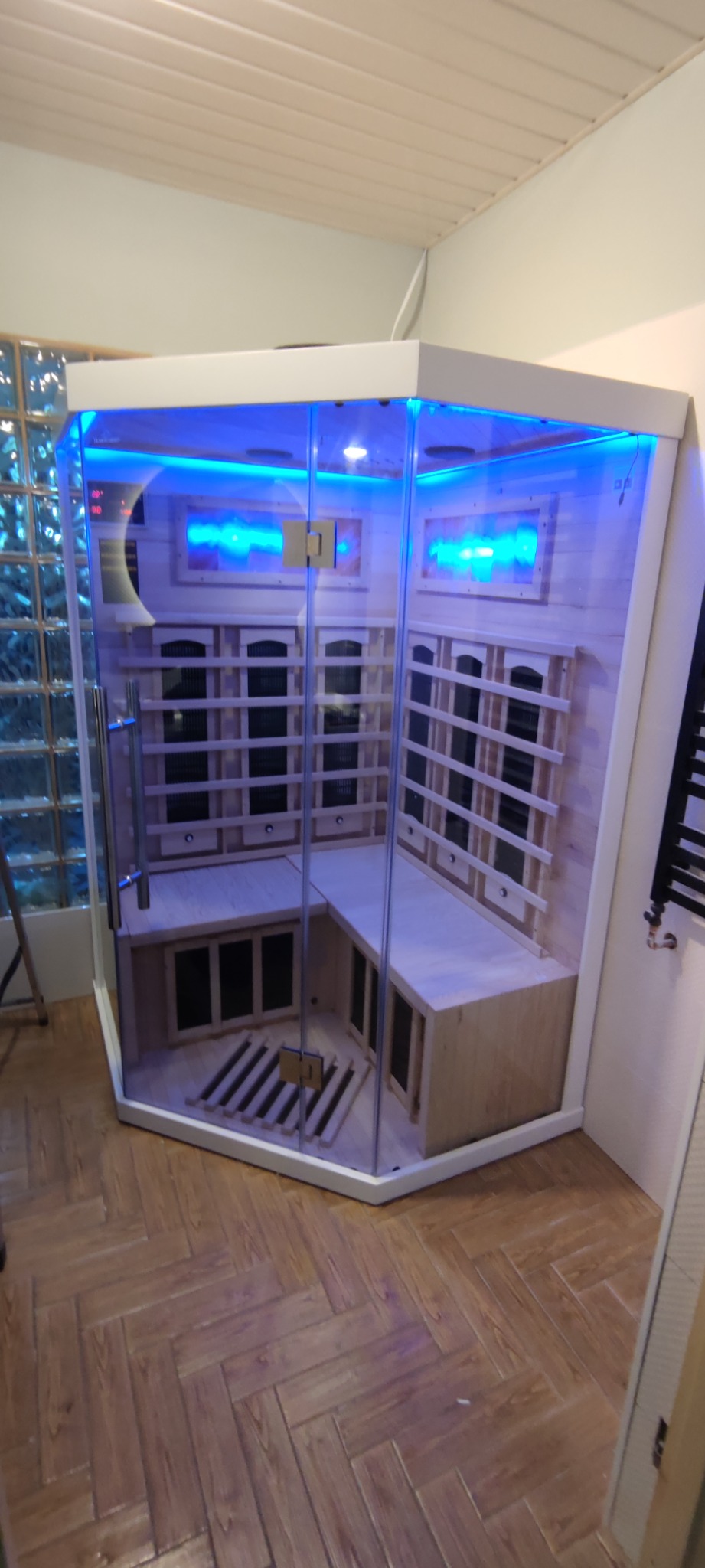 Gotowa kabina sauny infrared z drewnianymi ławkami i niebieskim oświetleniem LED w narożniku łazienki z przeszkloną ścianą z luksferów.