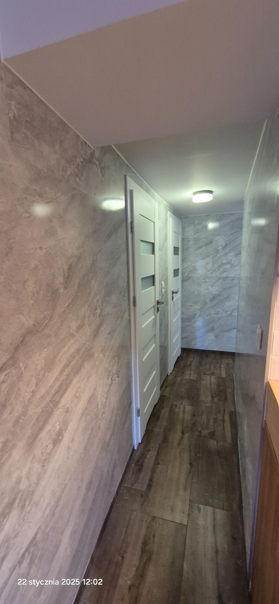 Korytarz wykończony płytkami imitującymi marmur na ścianach i drewnopodobnymi na podłodze, dwie białe drzwi z przeszkleniami, oświetlenie sufitowe.