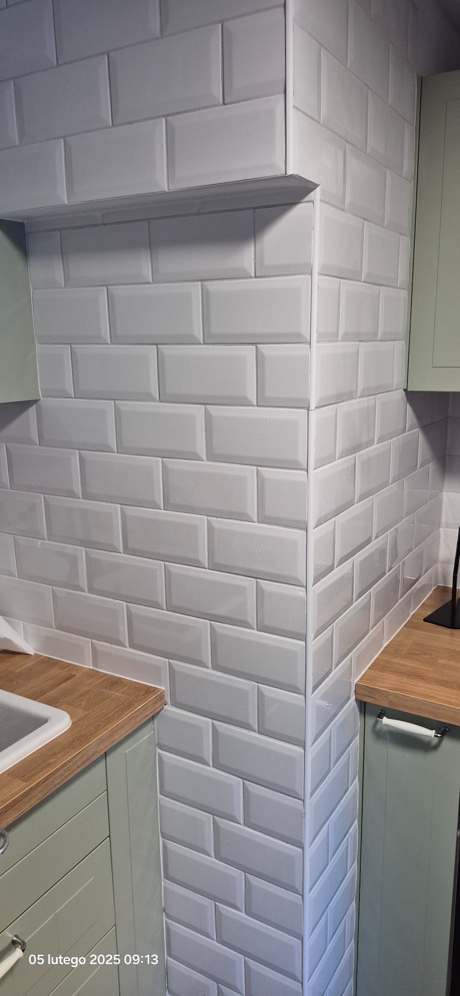 Wykończona ściana kuchni z białymi, prostokątnymi płytkami ułożonymi w cegiełkę, widoczny fragment blatu z jasnego drewna oraz szafki w odcieniu pistacjowej zieleni.
