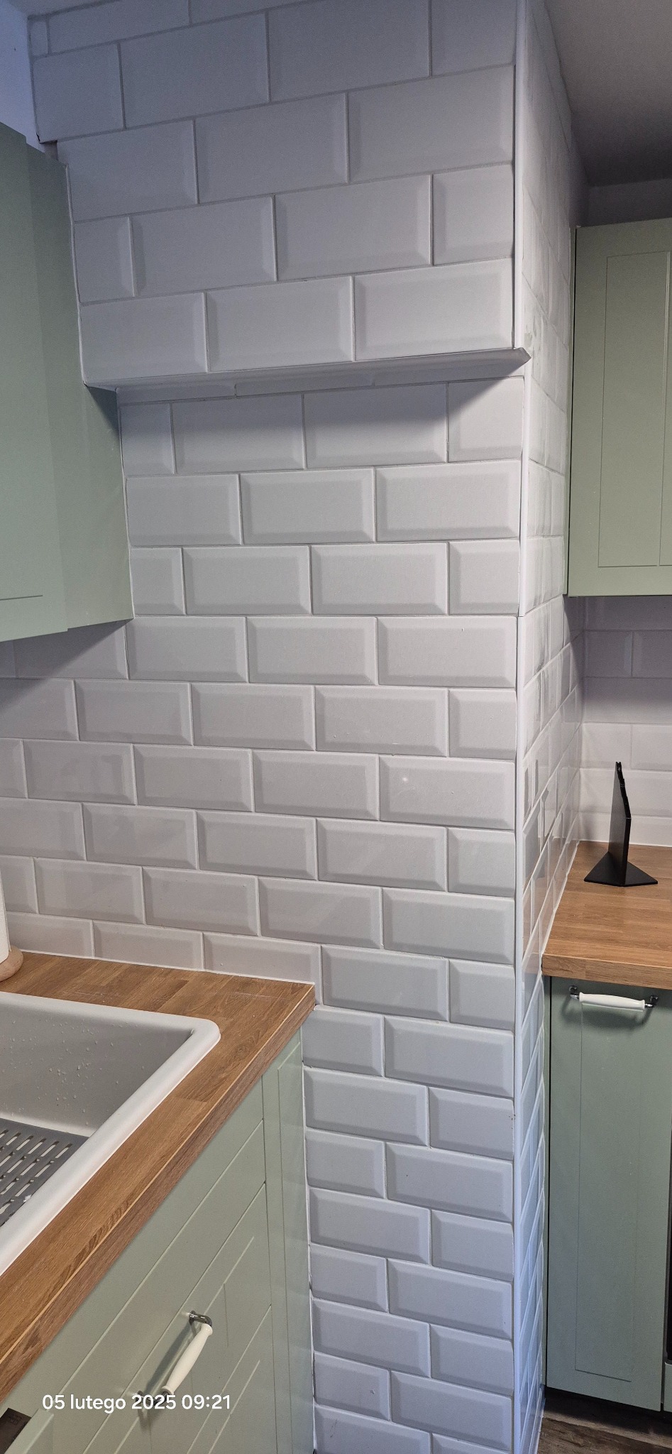 Fragment kuchni z białymi, błyszczącymi płytkami typu metro na ścianie, drewnianym blatem i jasnozielonymi frontami szafek. Widoczny zlew i narożnik ściany.