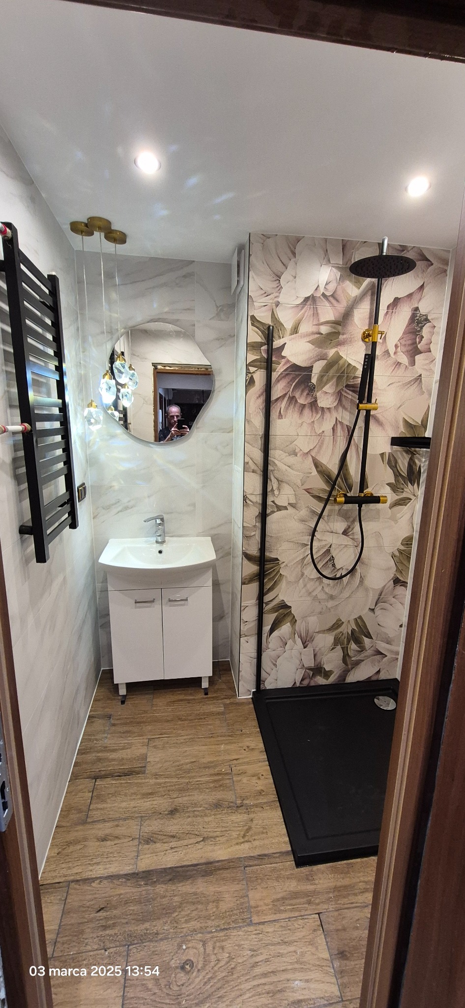 Nowoczesna łazienka z kabiną prysznicową typu walk-in, umywalką z szafką, grzejnikiem drabinkowym i lustrem o nieregularnym kształcie. Ściany wyłożone płytkami imitującymi marmur oraz płytkami...