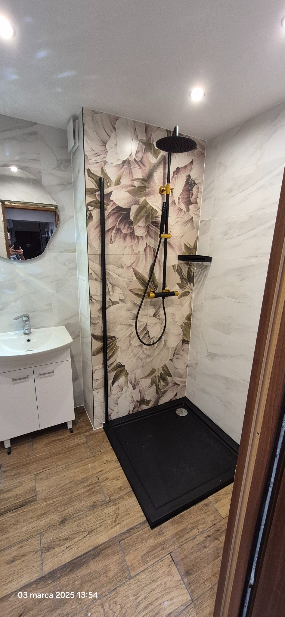 Nowoczesna łazienka po renowacji z kabiną prysznicową typu walk-in z czarnym brodzikiem i armaturą w kolorze czarnym i złotym. Ściany wyłożone płytkami imitującymi marmur oraz z motywem kwiatowym.