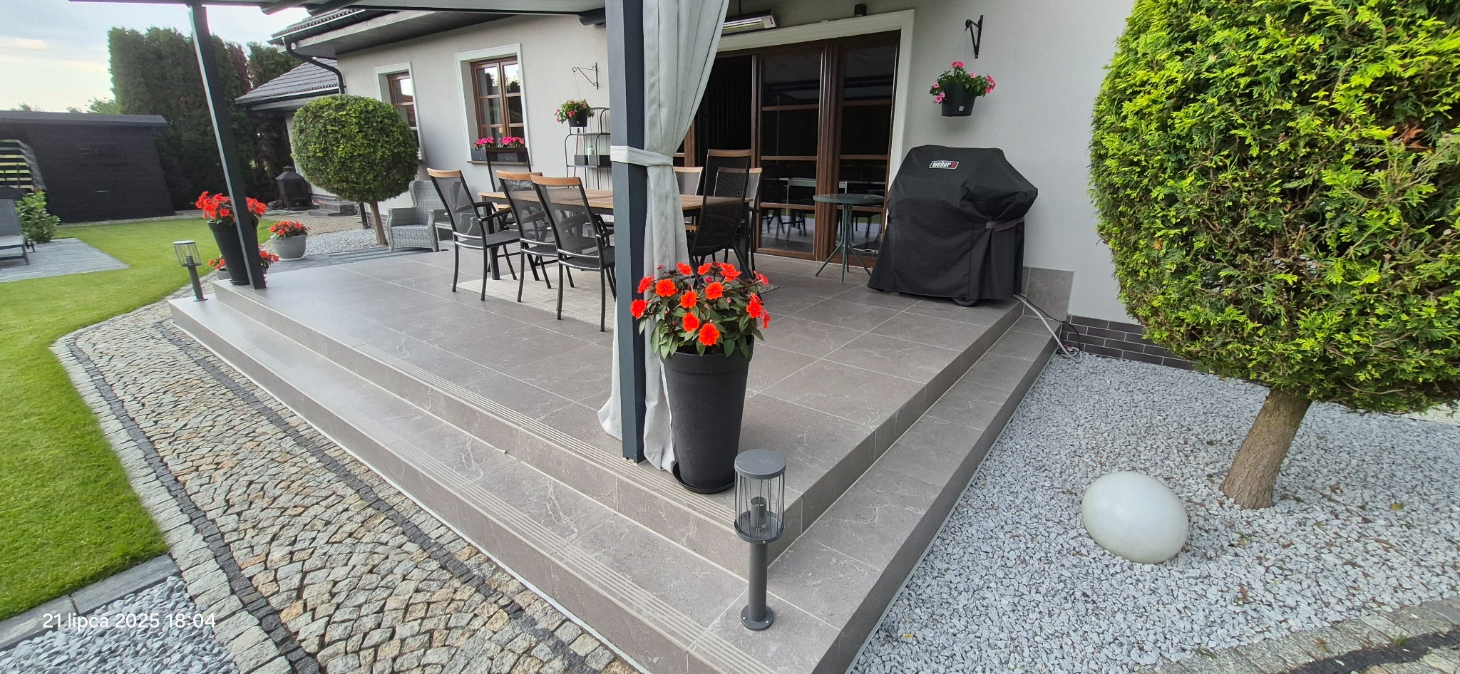 Wykończony taras z szarych płytek, schodami i obrzeżem z kostki. Na tarasie stół z krzesłami, grill i kwiaty w donicach. Oświetlenie tarasowe.