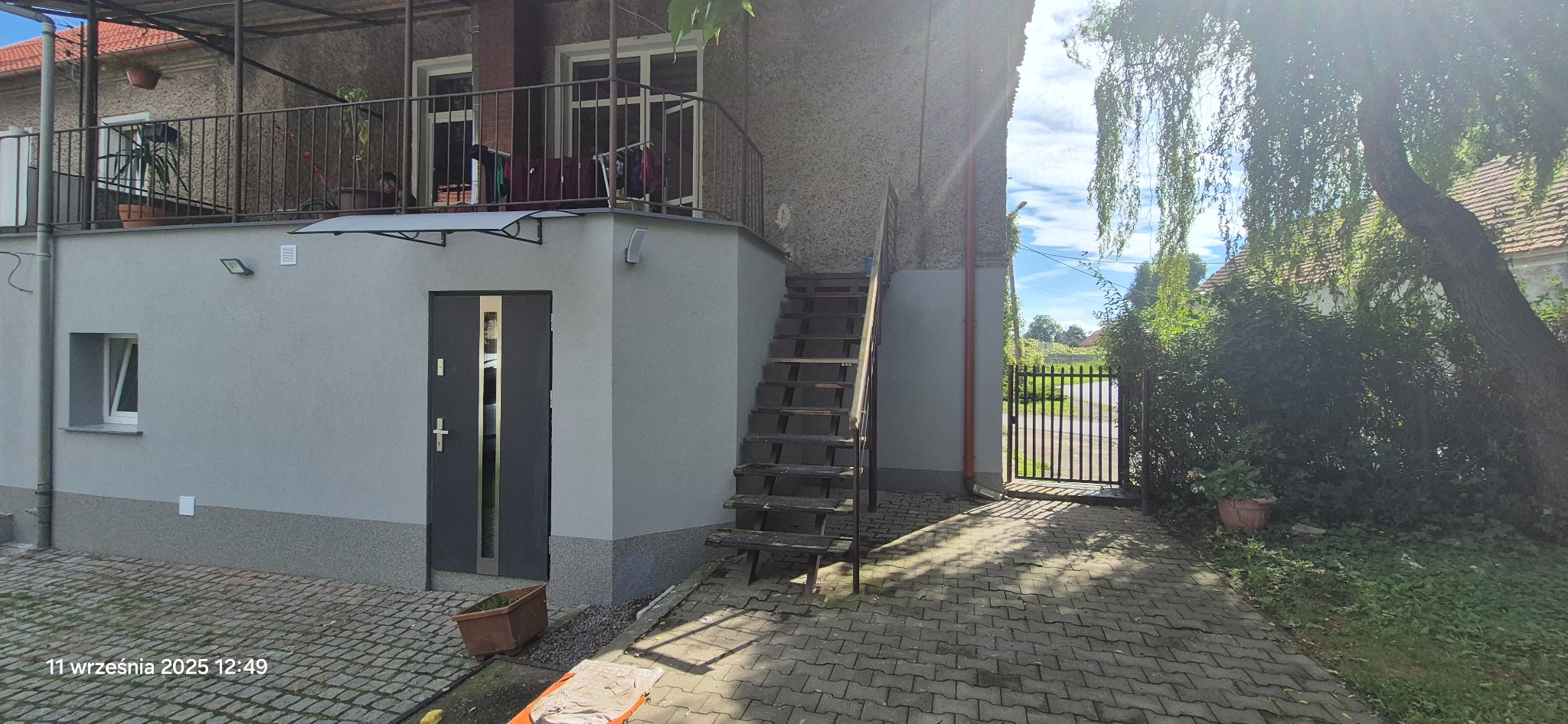 Szara elewacja domu z zewnętrznymi schodami prowadzącymi na balkon. Widoczne okno, drzwi wejściowe i brama wjazdowa. Uwagę przyciąga kontrast między nową elewacją a starszą konstrukcją.