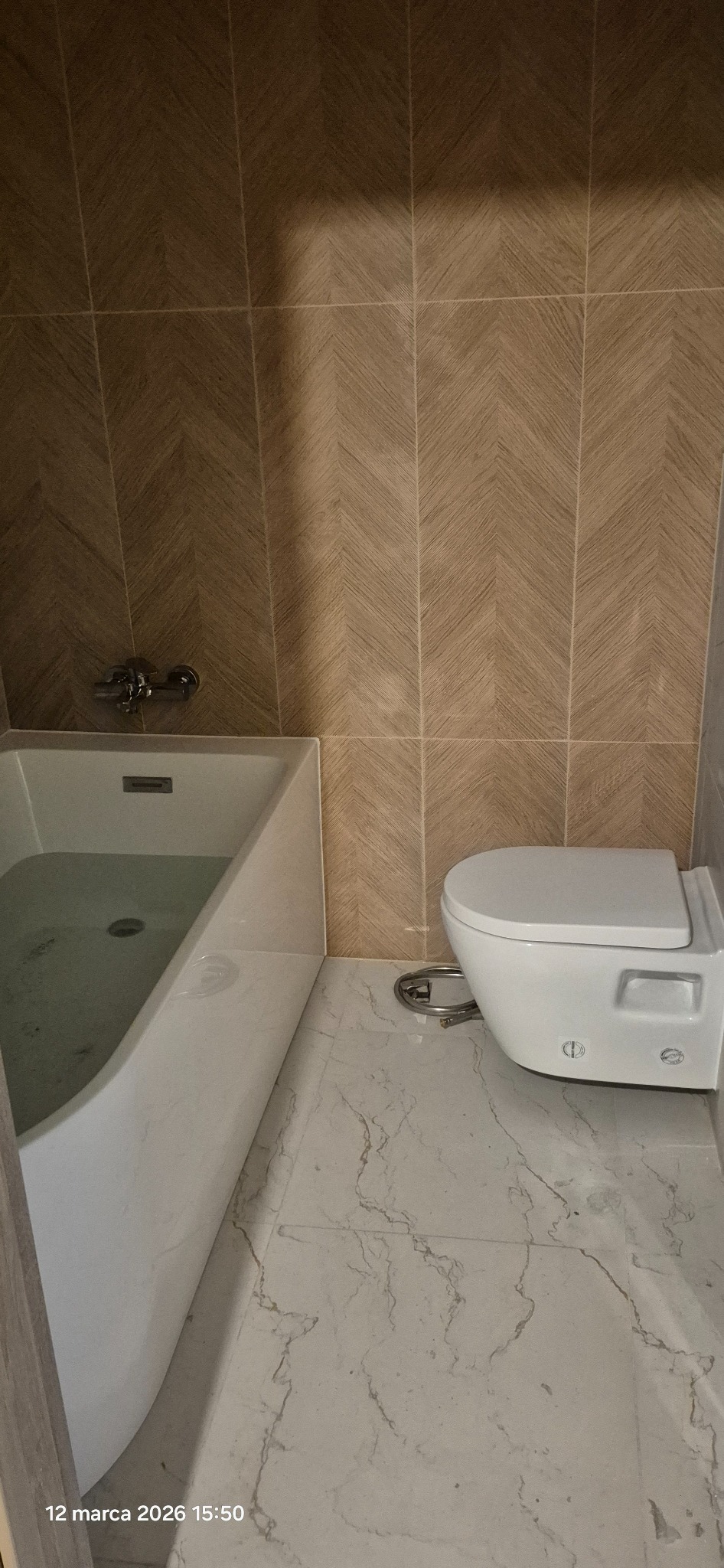 Wykończona łazienka z białą wanną wypełnioną wodą, podwieszaną toaletą i brązowymi płytkami ułożonymi w jodełkę na ścianie oraz marmurową podłogą.