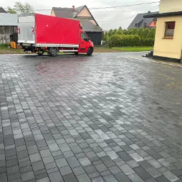 Ułożona, szara kostka brukowa na posesji z zaparkowanym czerwonym samochodem dostawczym. Widoczne budynki w tle i świeżo ułożone obrzeża.