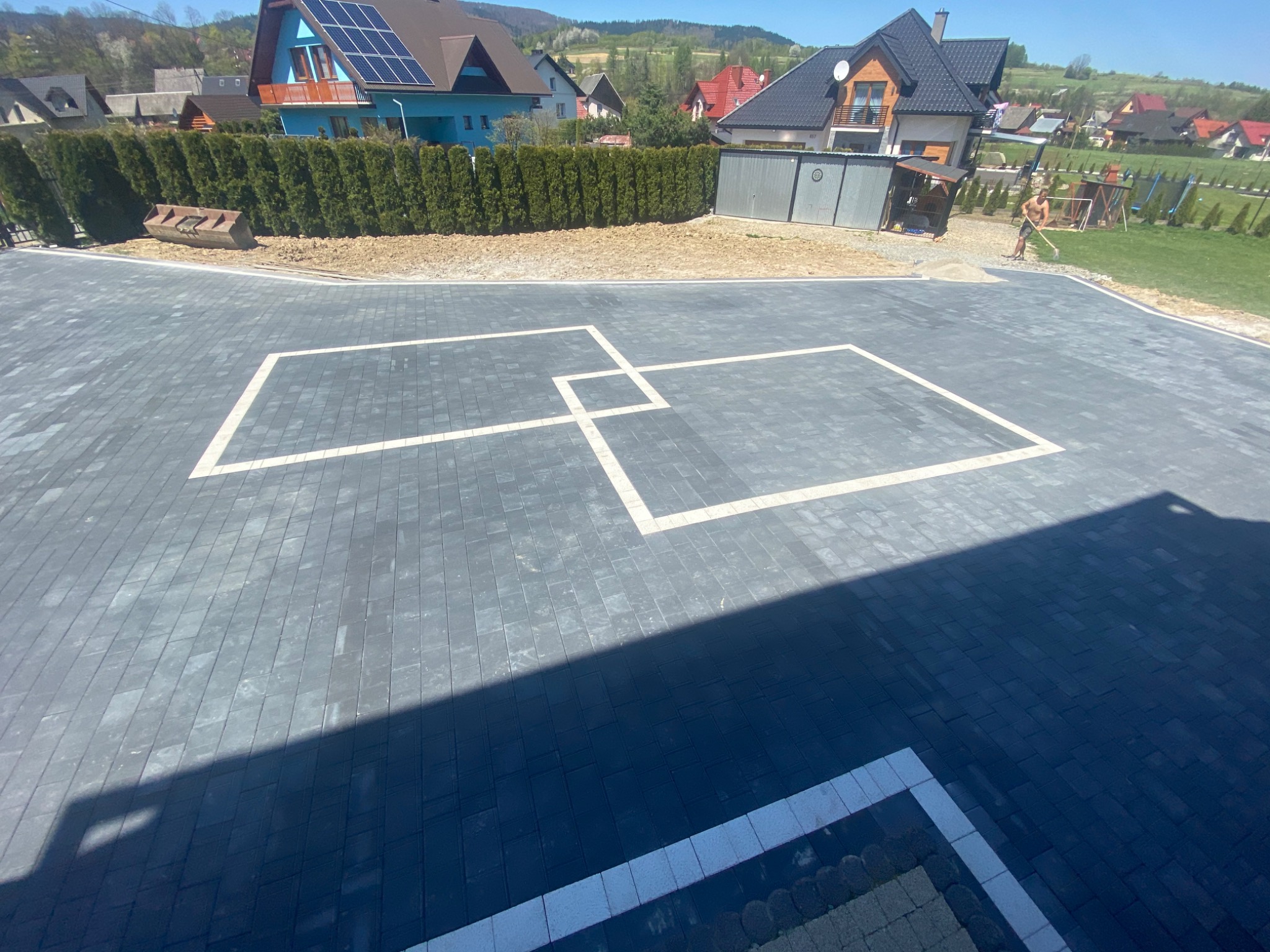 Ułożona kostka brukowa w Spytkowicach z dekoracyjnym wzorem z jasnych obramowań. W tle domy i zieleń, mężczyzna wyrównuje teren. Widok z góry, cień.