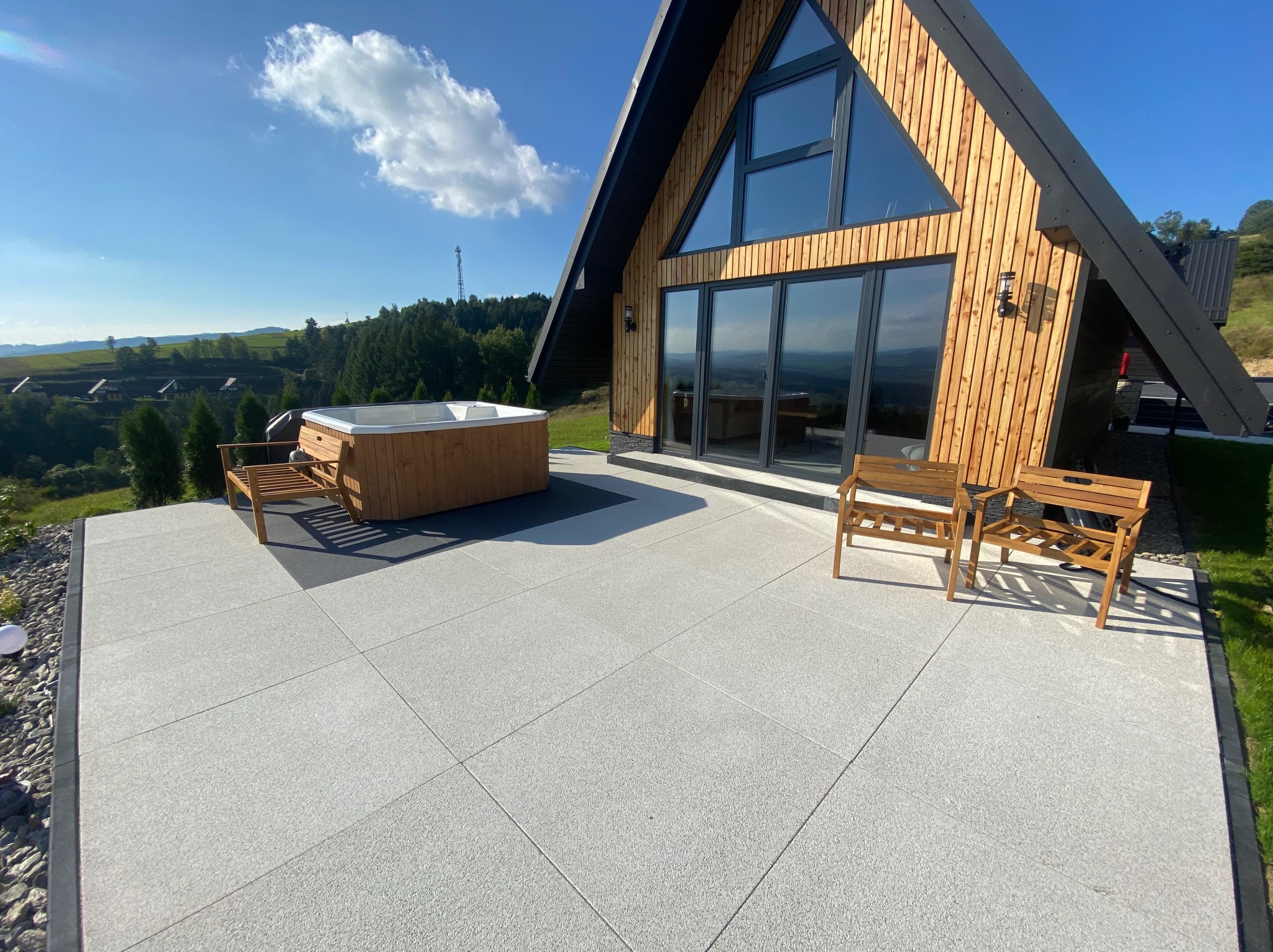 Nowoczesny taras z jasnoszarej kostki brukowej, jacuzzi i drewnianymi ławkami przed domem w stylu A-frame z widokiem na zielone wzgórza w słoneczny dzień.