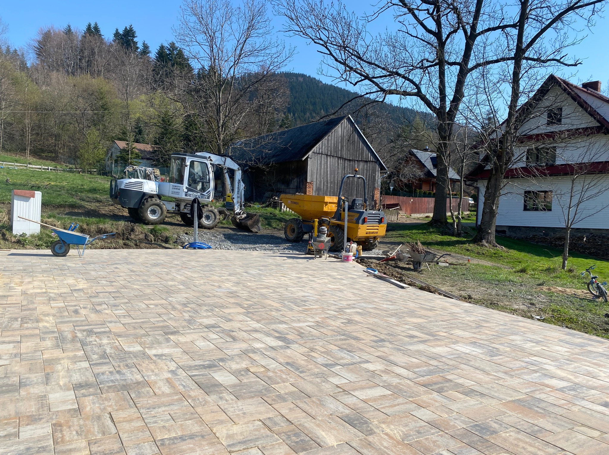 Ułożona kostka brukowa w odcieniach beżu i szarości na tle krajobrazu z maszynami budowlanymi i zabudowaniami. Widok na świeżo wybrukowany teren.
