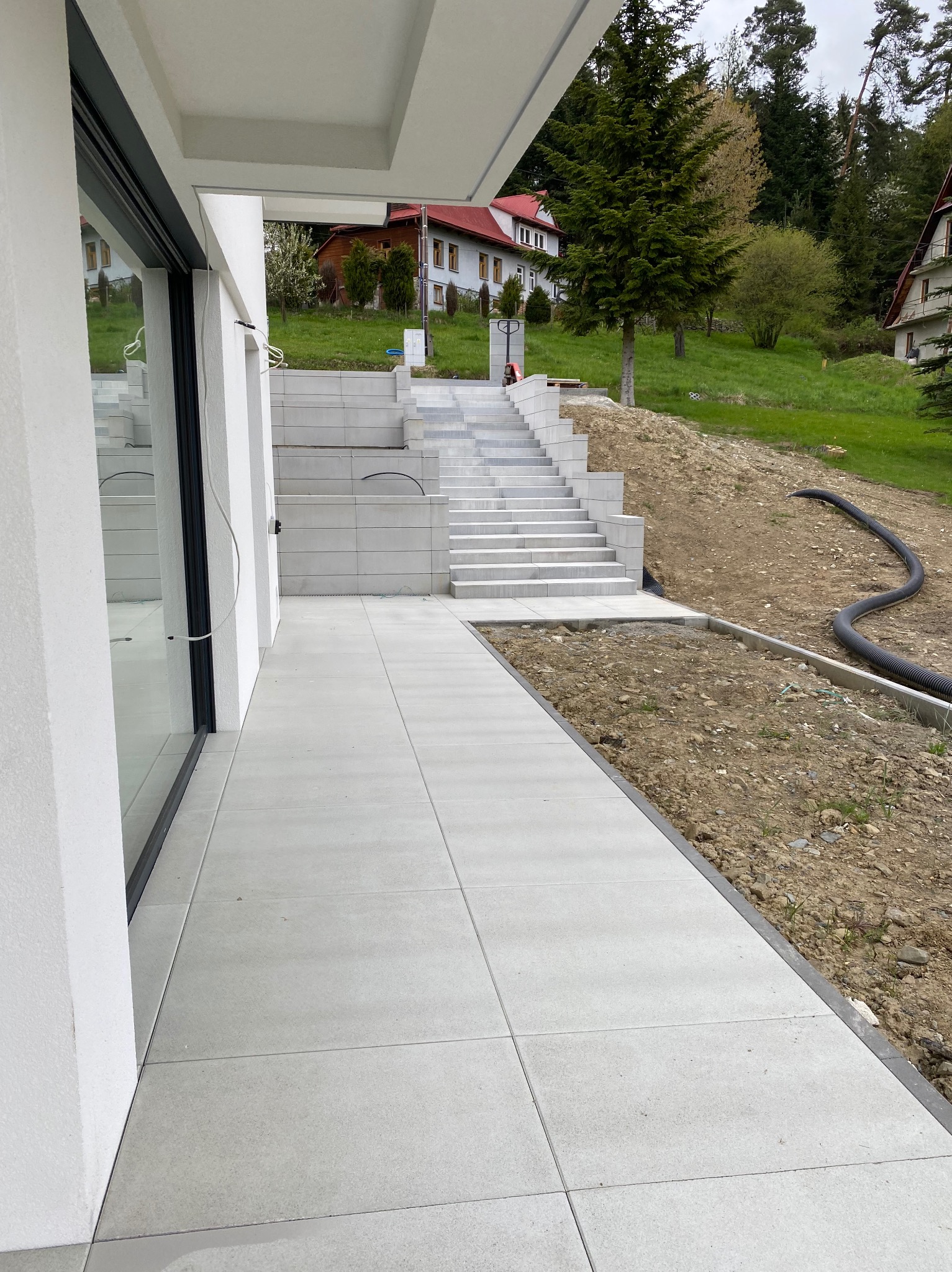 Nowoczesny taras z jasnych, kwadratowych płyt prowadzący do schodów z białych bloków. W tle domy na wzgórzu i fragment czarnego węża ogrodowego na ziemi.