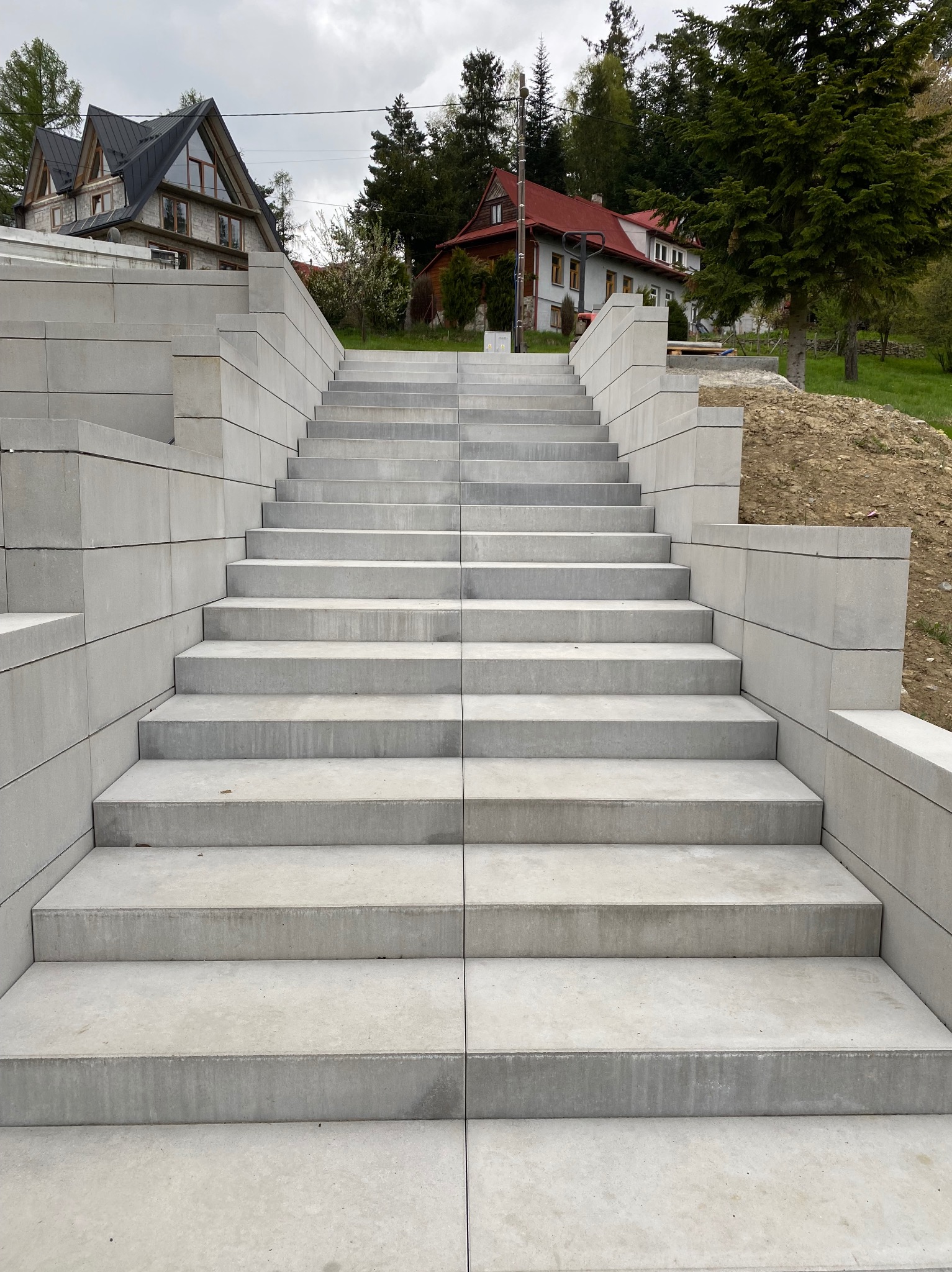 Nowoczesne schody zewnętrzne z szarej kostki brukowej prowadzące w górę, z widocznymi domami w tle. Symetryczny układ, minimalistyczny design.